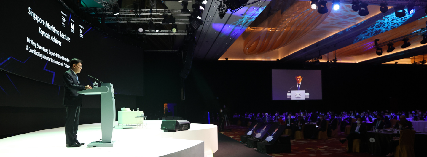 Man at podium onstage addressing audience. Screen displays lecture title. Stage lighting and ceiling art.